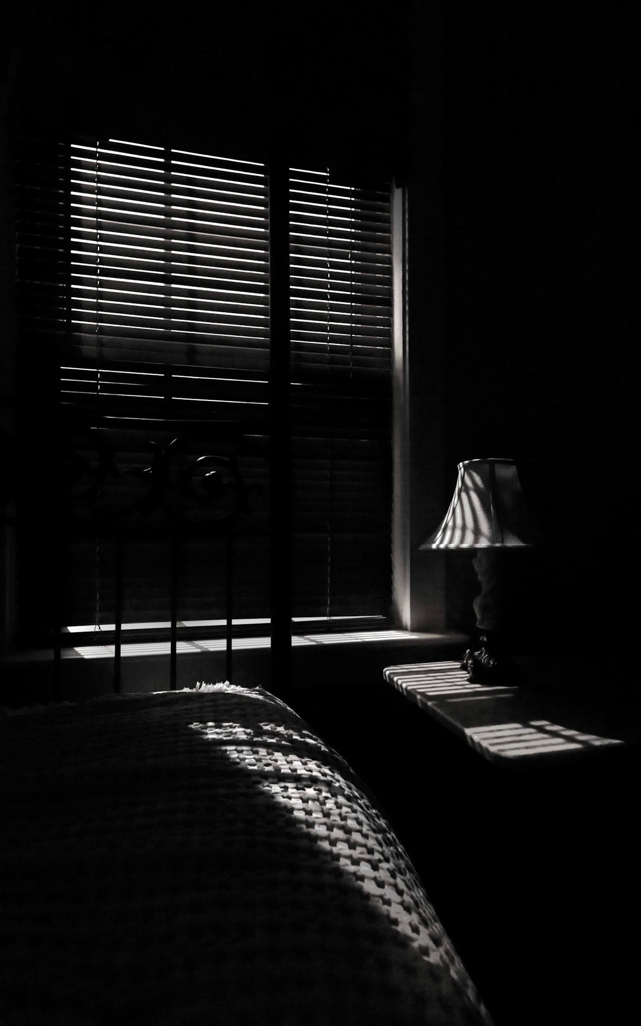 A black and white photograph of a window at night with a view of curtains and a lamp.