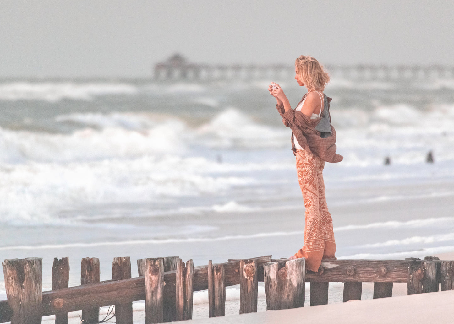 A woman in patterned flare pants stands on weathered wooden pier pilings at Naples, Florida beach, holding a phone to photograph the crashing white waves.