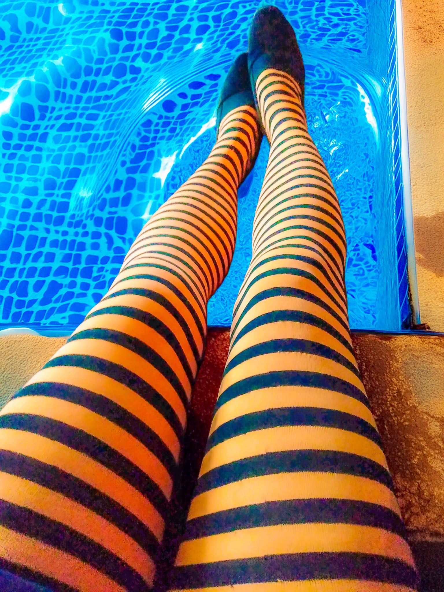High-contrast, top-down view of legs in bright orange and black striped stockings dangling over the edge of a vibrant blue swimming pool.