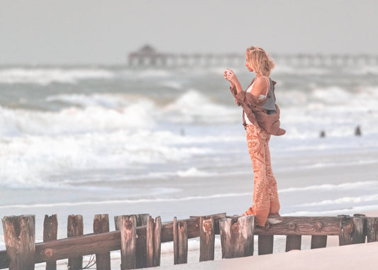 A woman in patterned flare pants stands on weathered wooden pier pilings at Naples, Florida beach, holding a phone to photograph the crashing white waves.